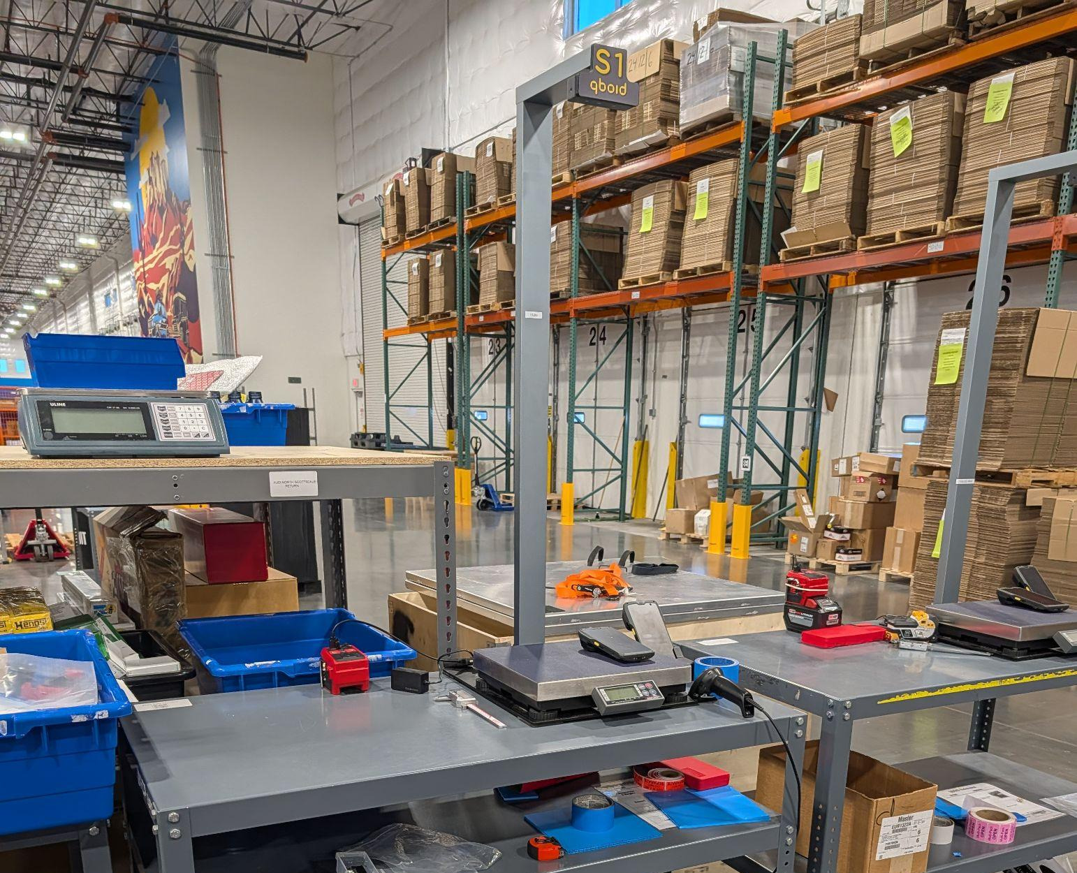 The S1 on a steel workbench in a warehouse receiving area: mast and overhead sensor above the scale, with pallet racking behind